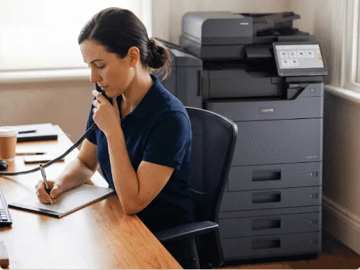 An account manager at a desk taking a call and writing notes, beside a Kyocera multifunction printer