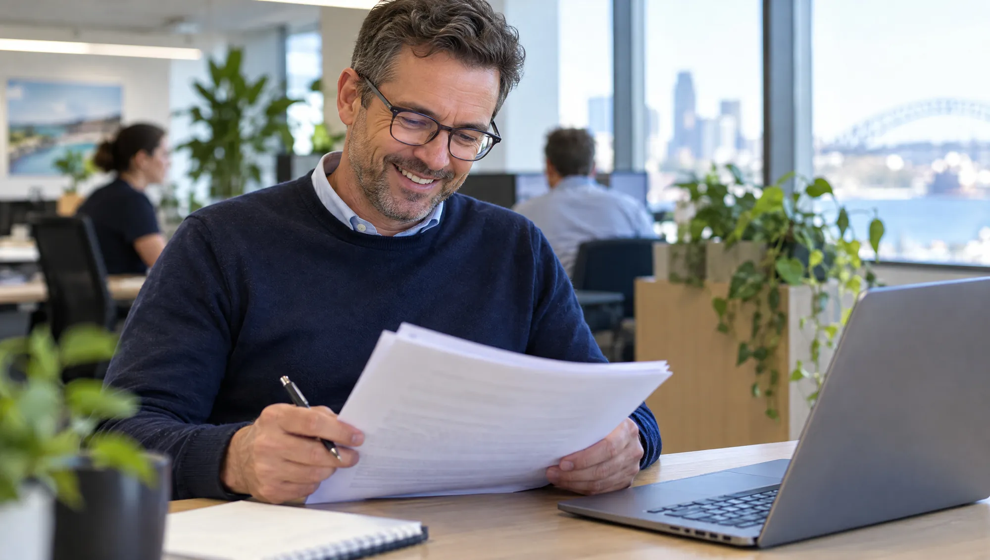 A smiling manager in glasses reads a stapled document at his desk, pen in hand.
