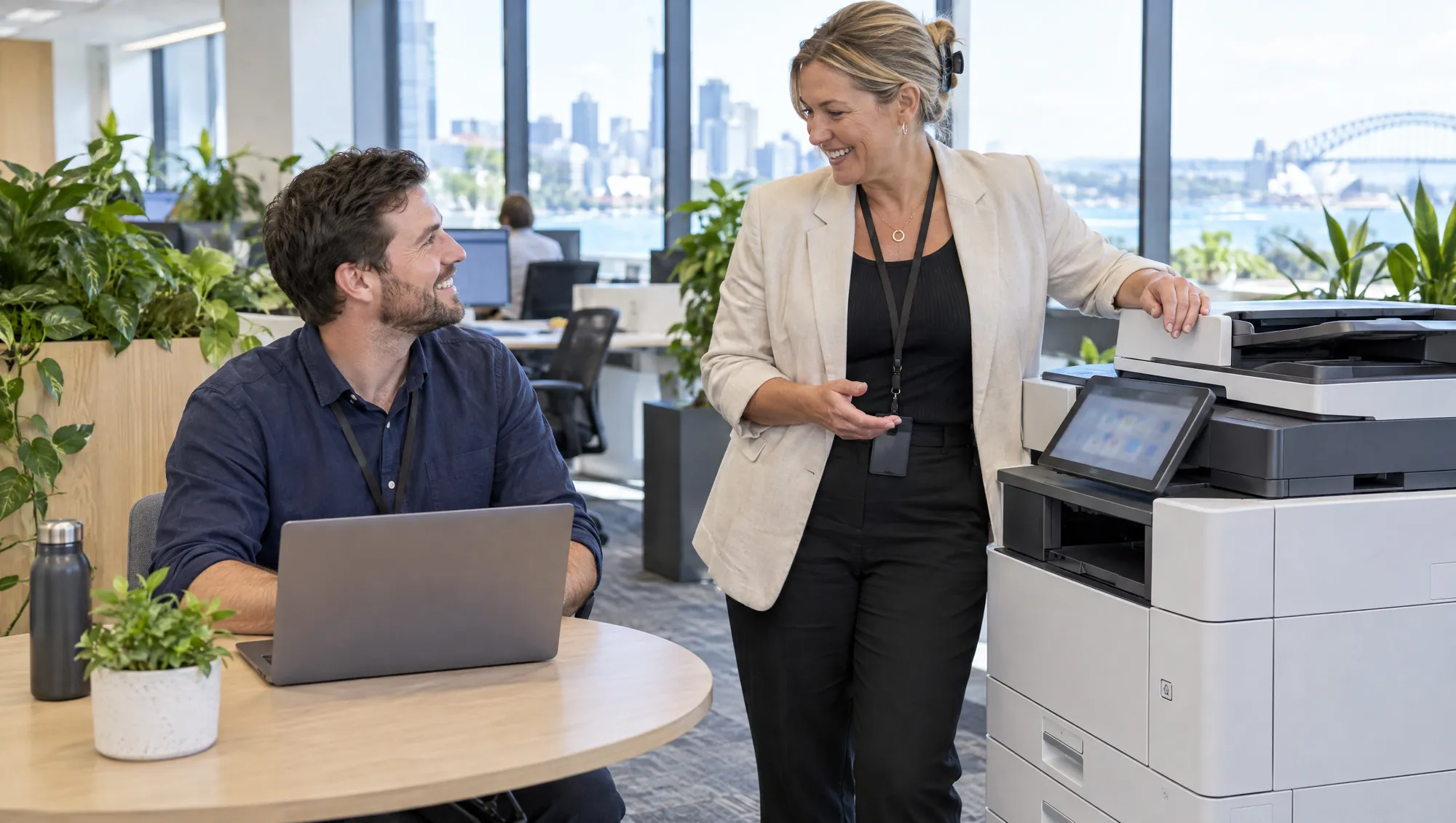 A manager at a laptop talks with a smiling colleague standing beside a large office printer.