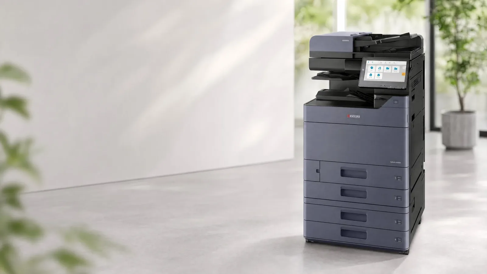 A Kyocera multifunction printer standing in a bright empty office, daylight from a window wall on the right and plants behind it