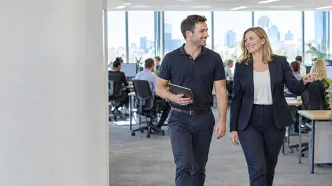 A consultant with a tablet walking an open plan office floor with a smiling office manager who points across the room