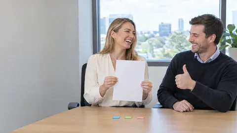 An accounts officer holding up one blank sheet and laughing with a colleague giving a thumbs up, three coloured tabs on the desk