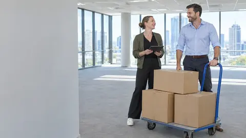 A facilities manager and a colleague laughing beside three plain moving boxes on a trolley in a bright empty office