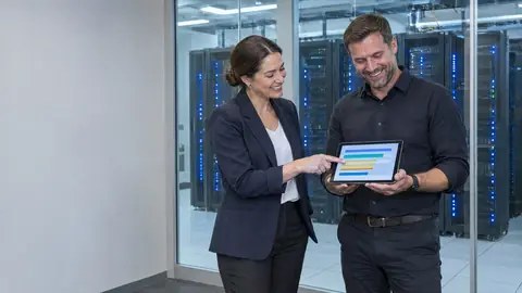 An IT manager and a colleague smiling over a tablet showing coloured bars, in front of a glass walled server room