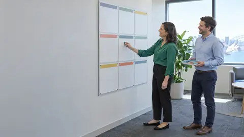 An office manager placing a magnet on one of twelve blank month panels while a laughing colleague holds the rest
