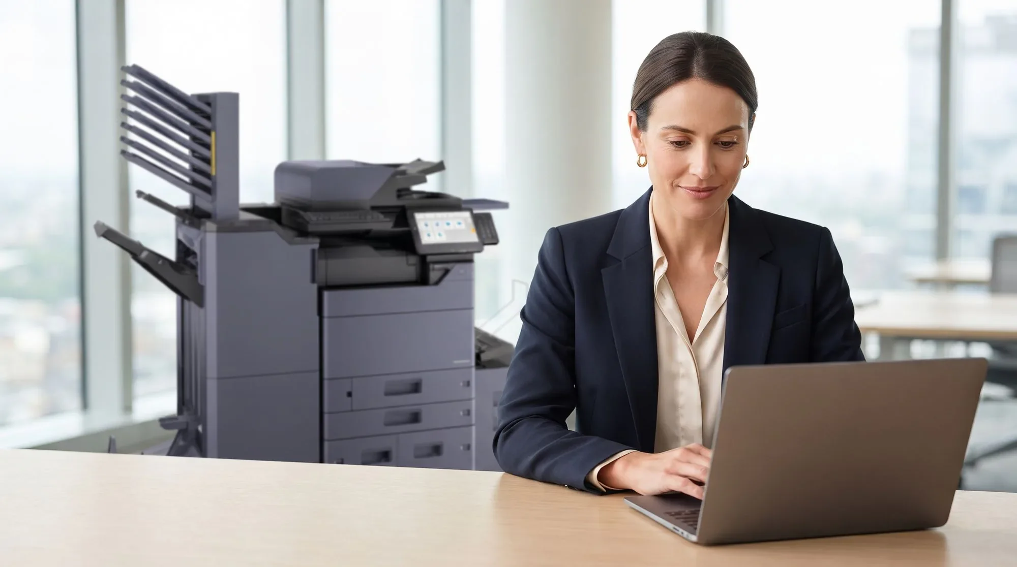 A woman working on a laptop at her desk, with a Kyocera multifunction printer and finisher standing behind her by the windows