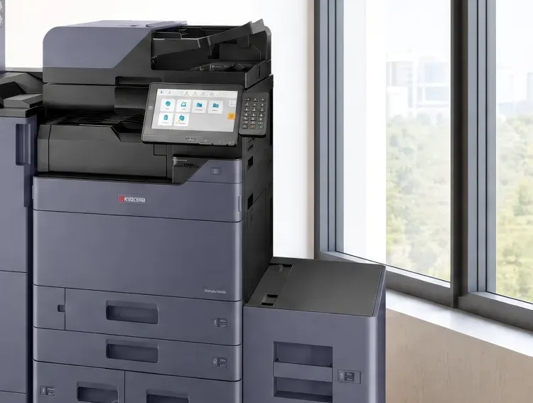 The control panel of a Kyocera multifunction printer at standing height, with the paper drawers below it